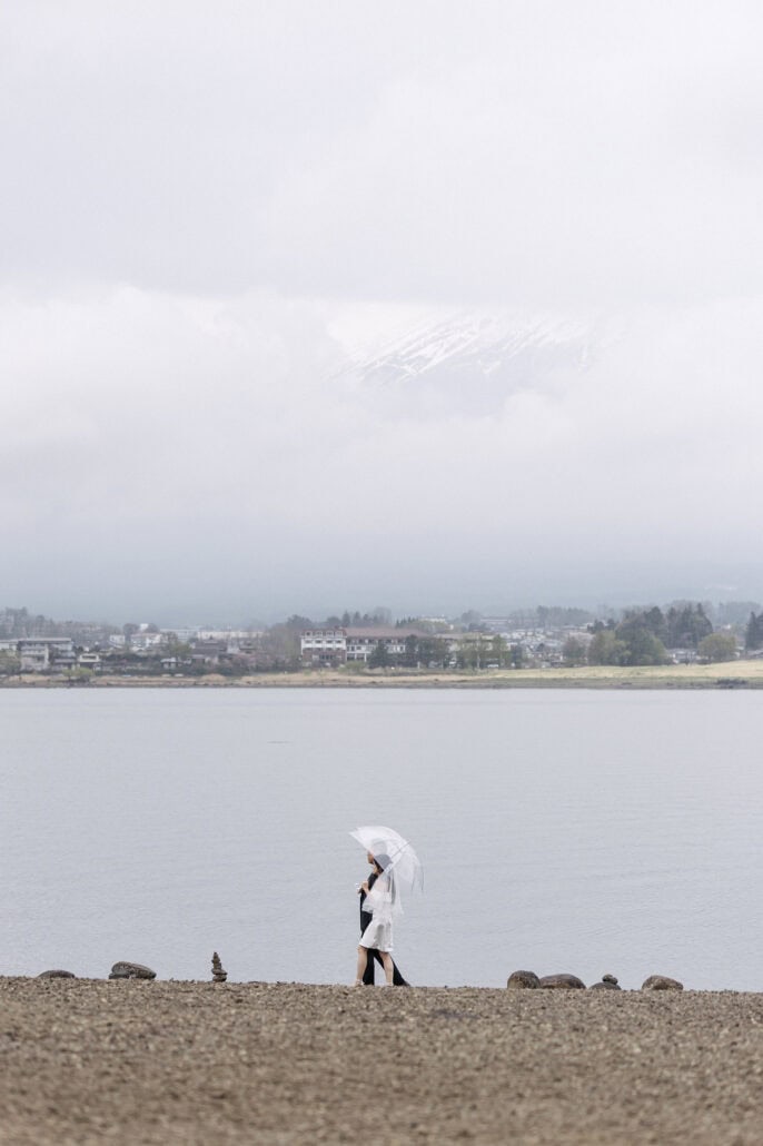 The couple walk the stony shore of Lake Kawaguchi under one clear umbrella, the water flat and grey and the snow on Mt Fuji faint in cloud.