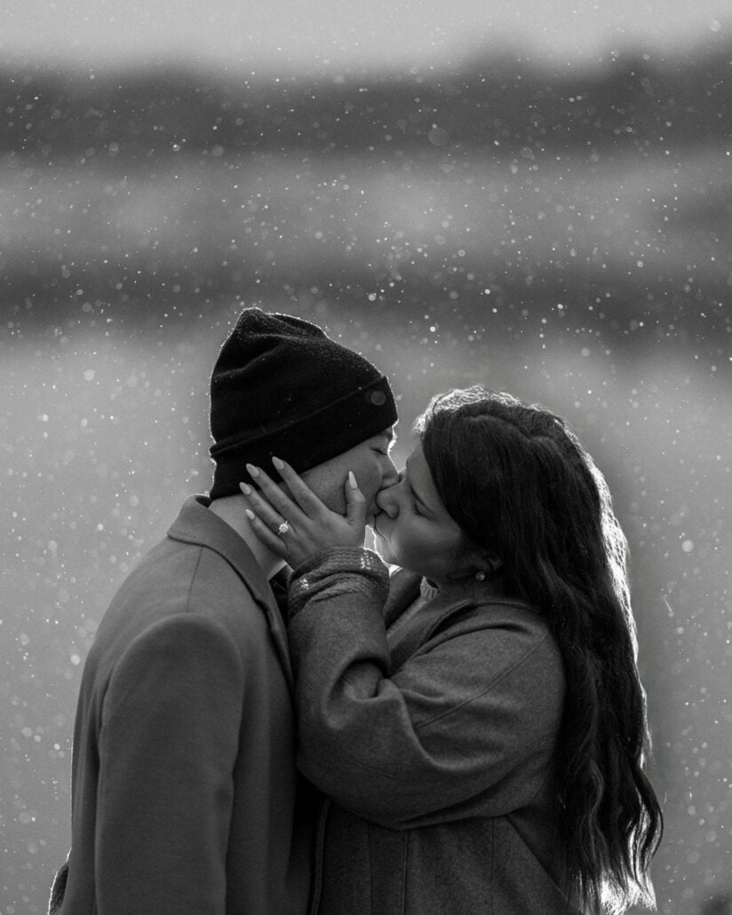 A close kiss as snowflakes fall around them, her ring hand on his face, in black and white after the proposal.