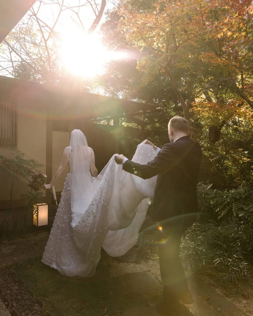 Komorebi, sunlight breaking through autumn leaves, flares behind the groom as he lifts the bride's train past a paper lantern in the ryokan garden.
