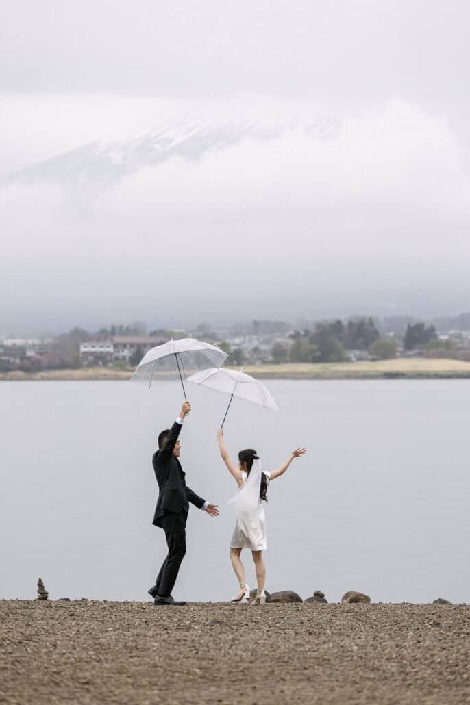 The bride and groom dance on the shore of Lake Kawaguchi with clear umbrellas held high, her free arm flung out and Mt Fuji faint in the cloud.