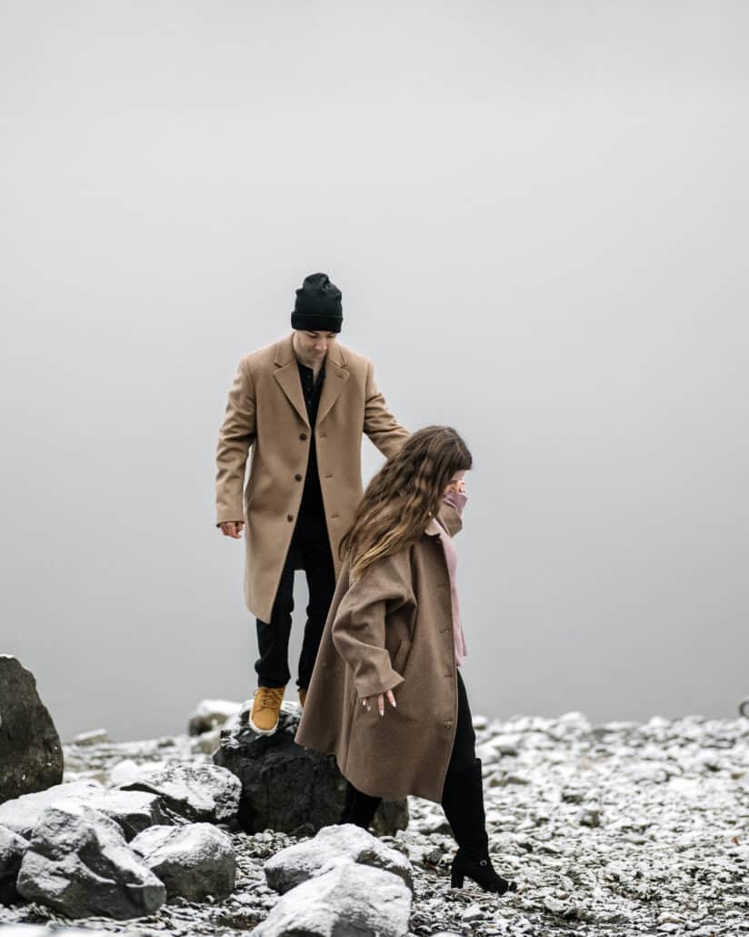The engaged couple step down over snowy rocks at the water's edge, laughing, hand in hand in their winter coats.