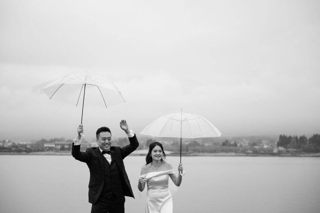In black and white, the bride and groom smile under clear umbrellas on the shore of Lake Kawaguchi, the groom's free hand raised in a wave.