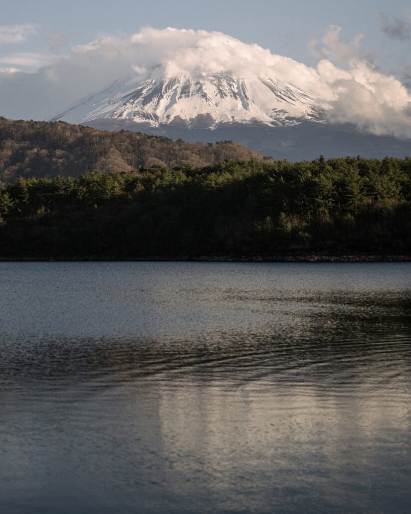 Mount Fuji, capped with cloud, rises above a forested ridge and a still lake, one of the Fuji Five Lakes, on a March afternoon.