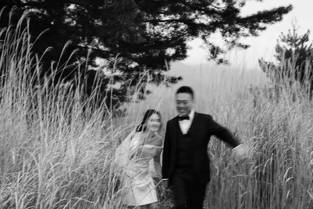 In black and white, the bride and groom hurry through tall dry grass under a pine near Lake Kawaguchi, smiling and blurred with movement.