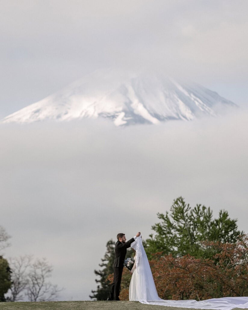 The groom lifts the bride's long veil on a lawn by Lake Kawaguchi as Mt Fuji rises out of a sea of clouds behind them.