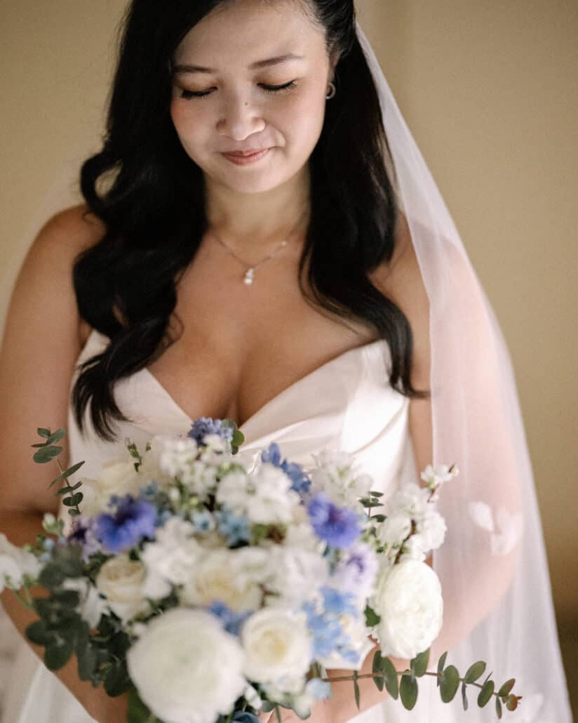 The bride looks down at her bouquet of white roses, blue cornflowers and eucalyptus, getting ready for her spring wedding in Izu.