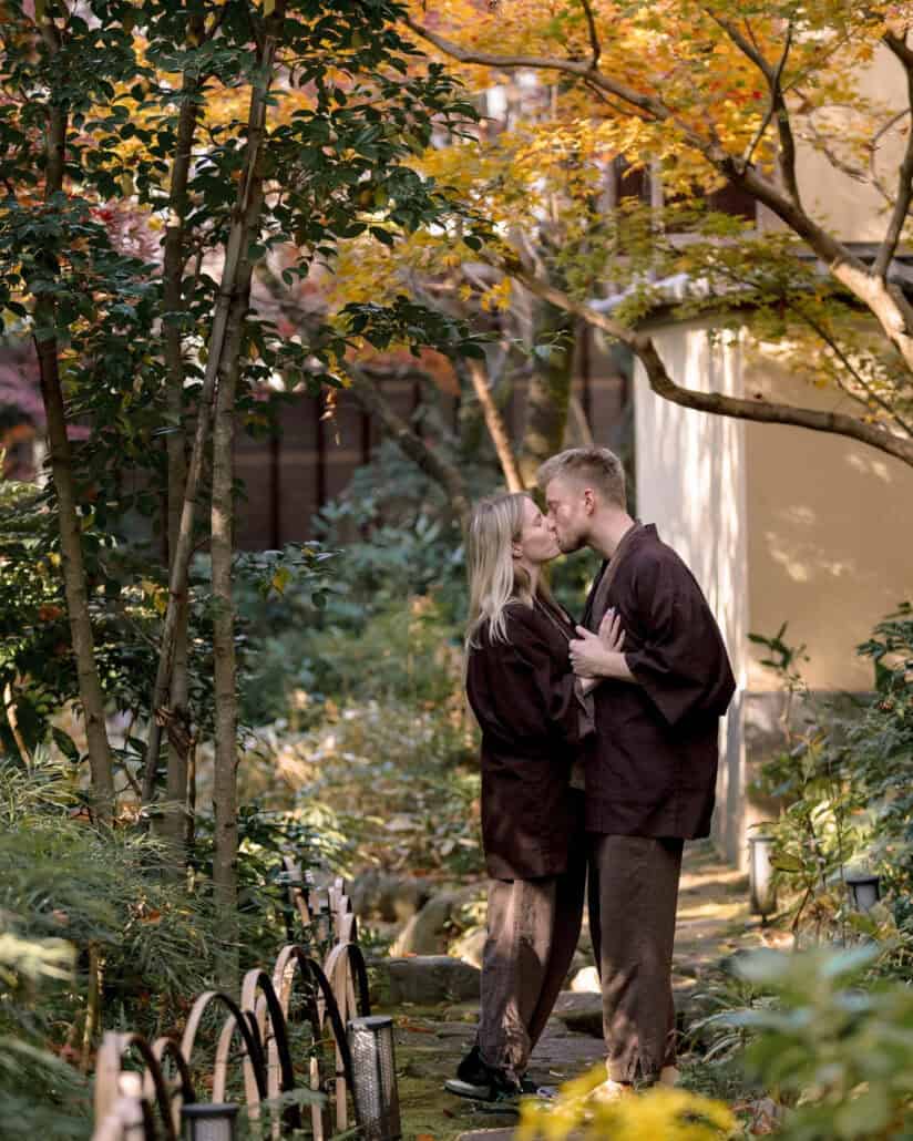 The couple kiss on a mossy stone path in matching brown samue, gold autumn leaves above, in the garden of the Hadano ryokan where they eloped.