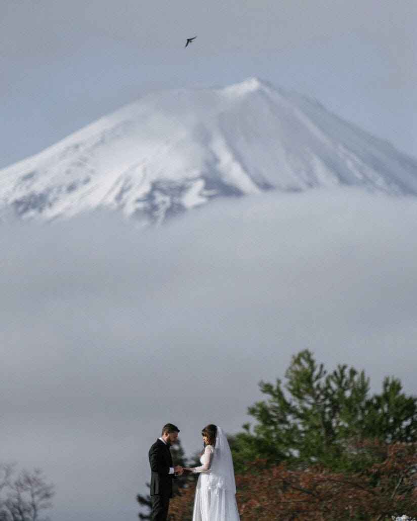 The groom slips a ring onto the bride's finger during their Kawaguchiko wedding vows, as the snowy summit of Mt Fuji rises out of the cloud above.