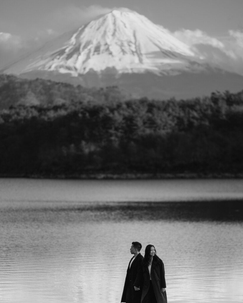 Mount Fuji rises above the forest and a still lake, the engaged couple walking small along the shore below, in black and white.