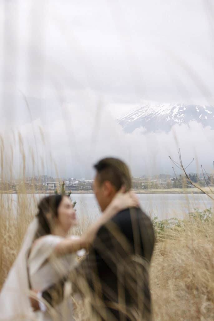 The bride wraps an arm around the groom's neck in tall dry grass, both soft in the foreground, the lake and Mt Fuji's snowfields sharp behind.