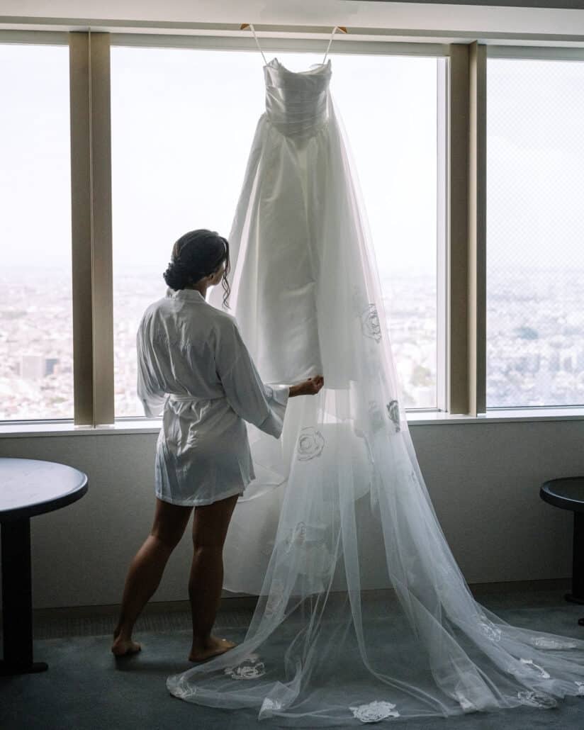 The bride, in a white robe, reaches up to her wedding dress hanging in a window high above Tokyo on her wedding morning.