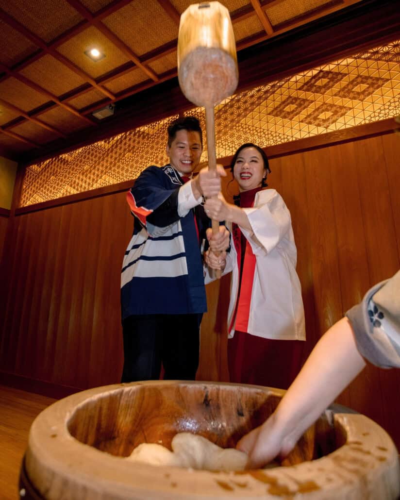 The bride and groom in happi coats lift a wooden mallet to pound mochi at their welcome party, a night their Jinya wedding film keeps.