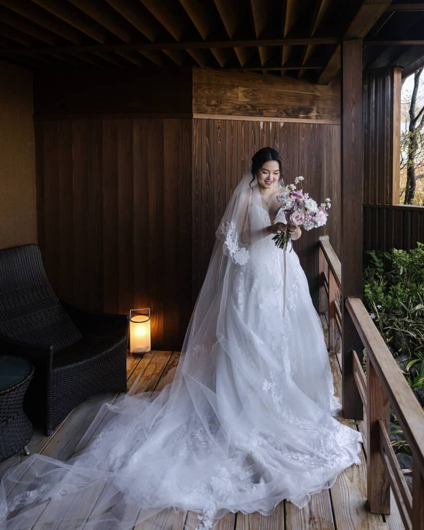The bride in her wedding dress and long veil holds a pink bouquet on the inn's wooden veranda, a lit lantern on the boards beside her.