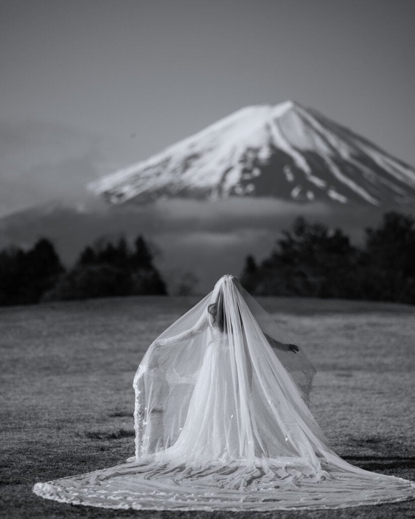 In black and white, the bride holds her long veil out across the lawn at Lake Kawaguchi, with Mt Fuji and a band of cloud ahead of her.