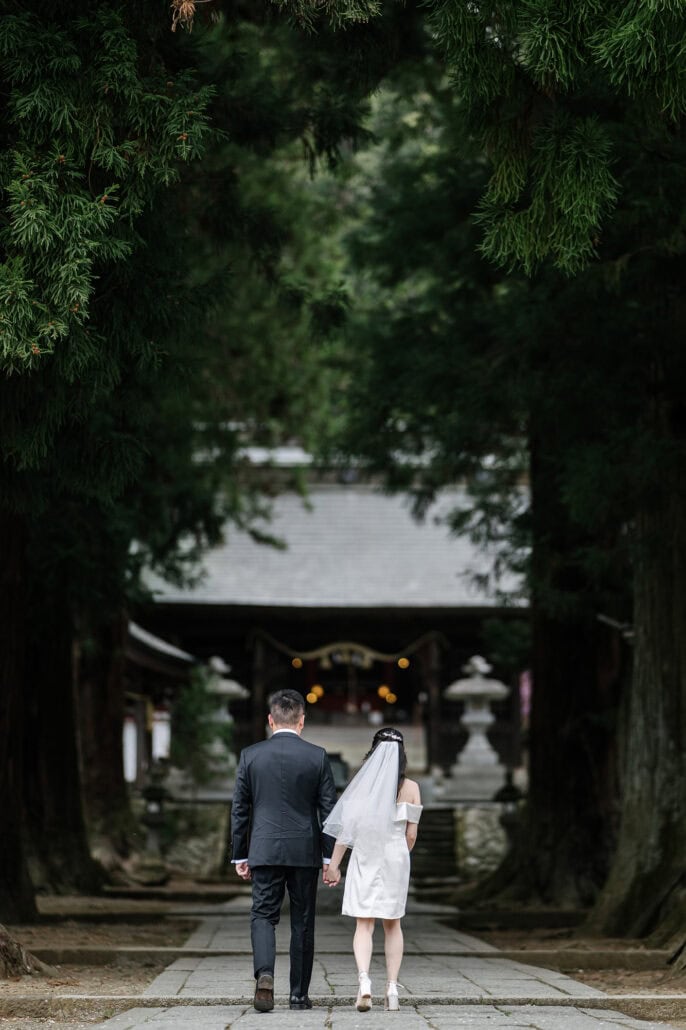 The couple walk hand in hand up a stone path between giant cedars toward a hall of Kawaguchi Asama Shrine, stone lanterns on either side.