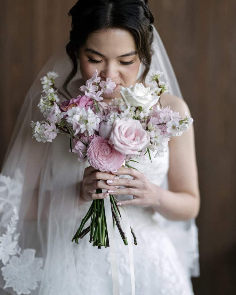 The bride holds her bouquet of pink and white roses and cherry blossom sprigs to her face on her destination wedding day in Japan.
