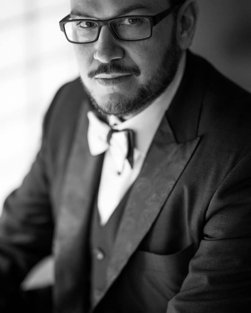 A black and white portrait of the groom in glasses, a bow tie and paisley lapels, in soft side light on the wedding morning in Shuzenji.