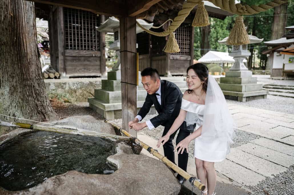 The bride laughs as she and the groom rinse their hands at the shrine's stone basin, fed by bamboo pipes, thick rope tassels hanging overhead.