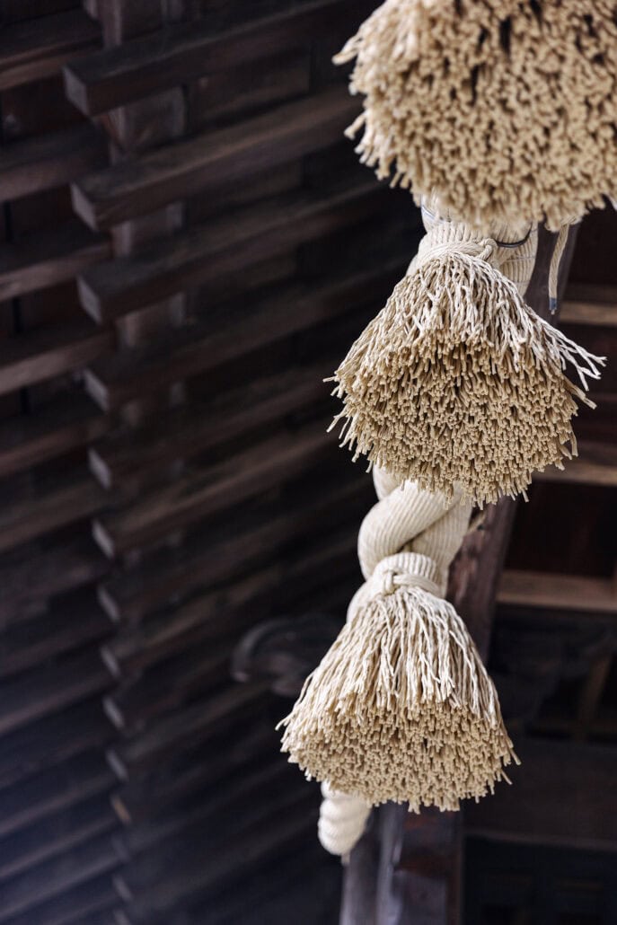 Pale tassels hang from a thick twisted rope against the dark wooden beams of a roof at Kawaguchi Asama Shrine, seen close up.