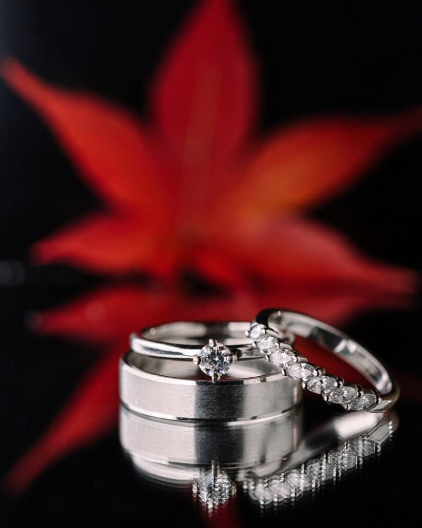 An engagement ring and two wedding bands reflect on a black surface in front of a blurred red maple leaf, a Japan elopement detail in autumn.