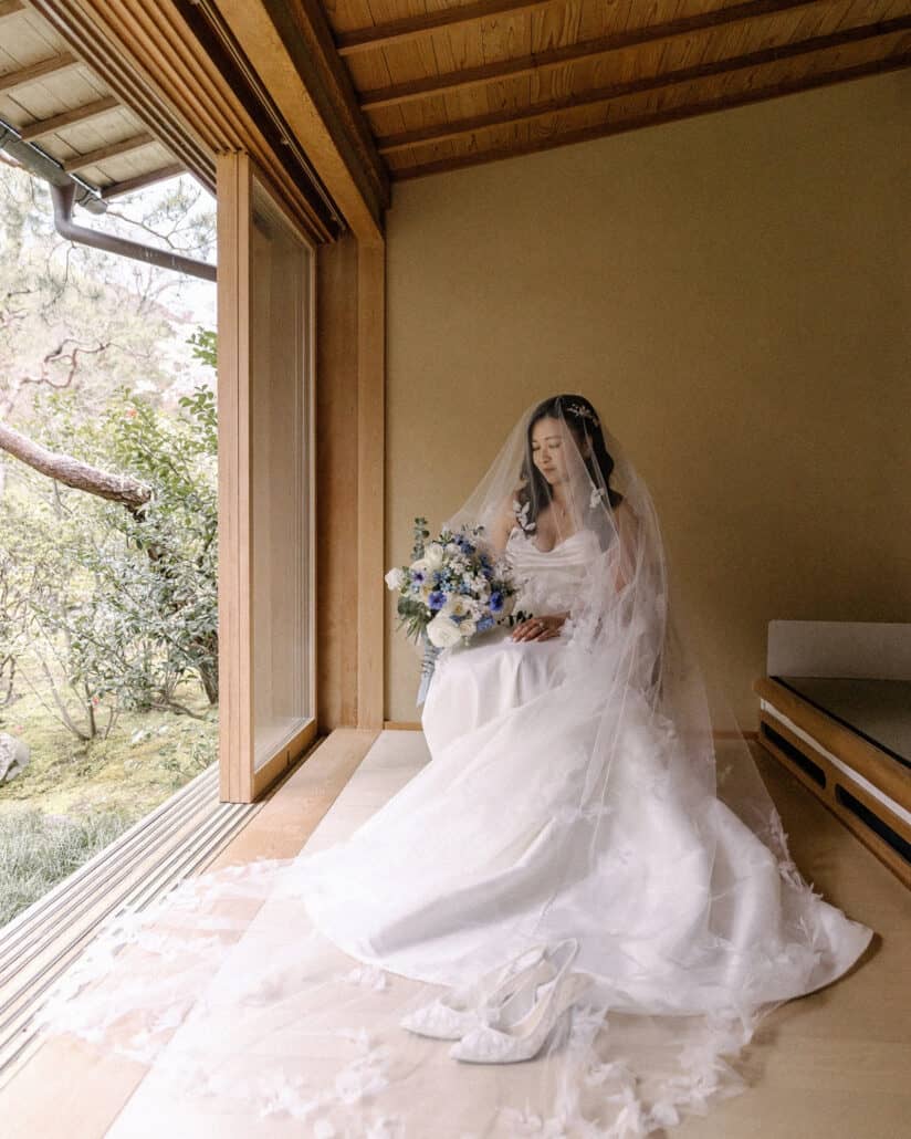 The bride sits under her veil with her blue and white bouquet by a garden window in her Asaba ryokan suite, her lace shoes at her feet.