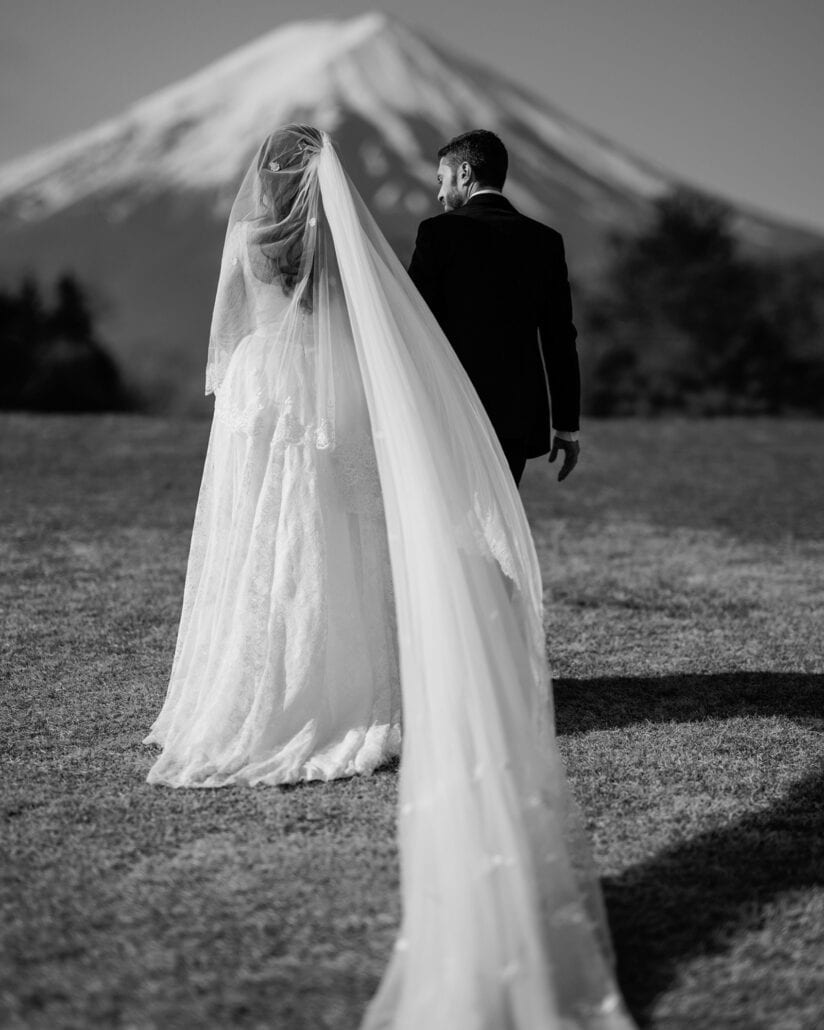 In black and white, the bride's long veil trails over the grass as the groom walks ahead toward snow-capped Mount Fuji on their elopement morning.