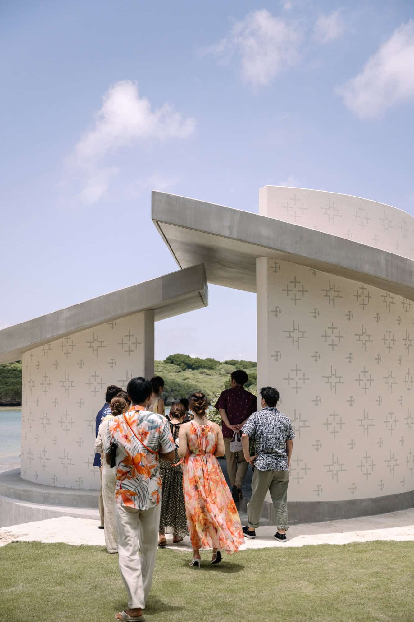 Guests in floral shirts walk into a round chapel beside the sea, a green headland across the water, at a resort wedding venue in Okinawa.