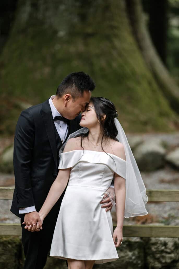 The groom rests his forehead against the bride's as they hold hands, her eyes closed, in front of the mossy trunk of a giant cedar at the shrine.