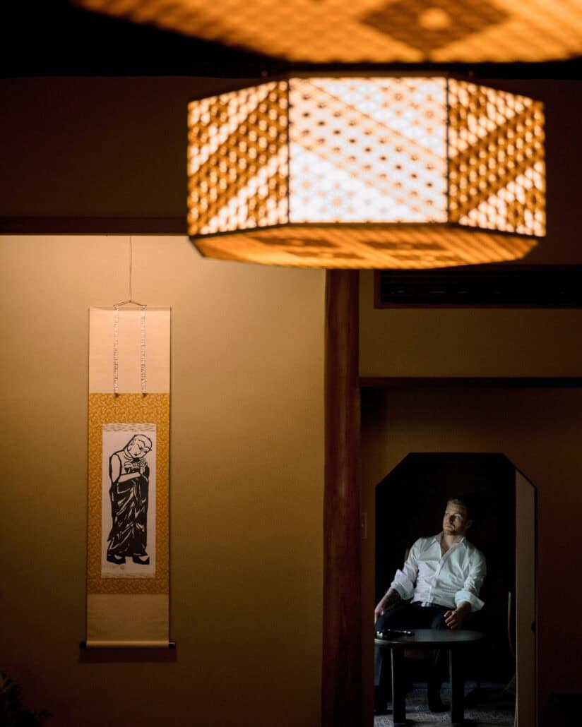 Past a lattice lantern and a hanging scroll, the groom sits in an open white shirt through a doorway, getting ready for a ryokan wedding in Japan.