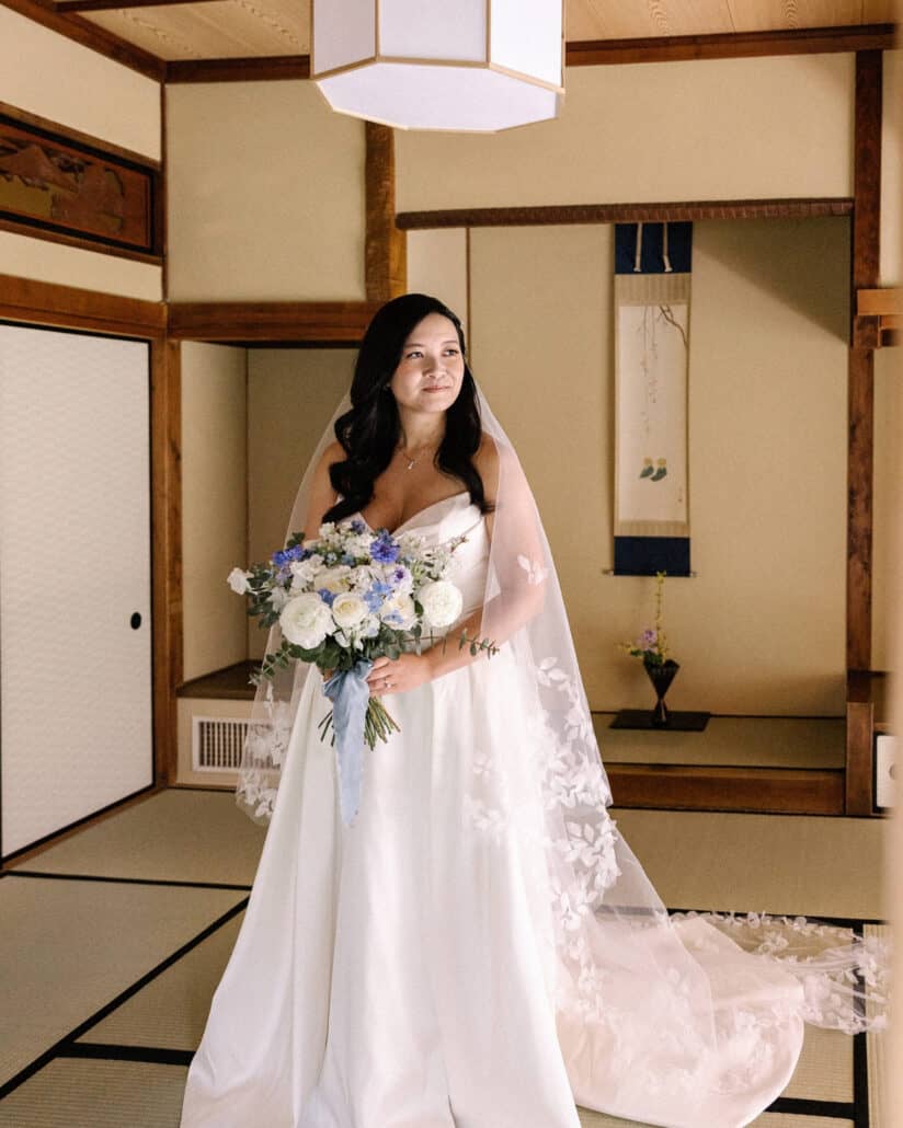 The bride stands in a tatami room at Asaba ryokan with her bouquet, her veil trailing on the mats, a hanging scroll and flowers in the alcove.