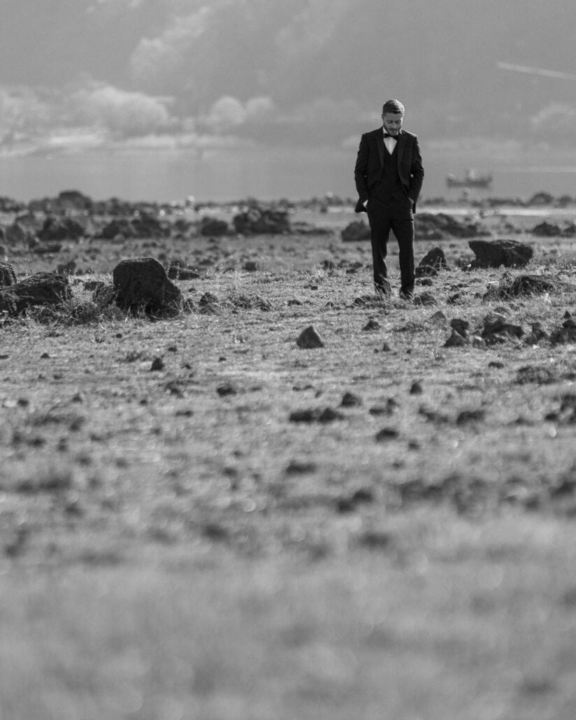 In black and white, the groom walks alone over the lava-rock shore of Lake Kawaguchi, hands in his pockets, a boat on the water behind him.