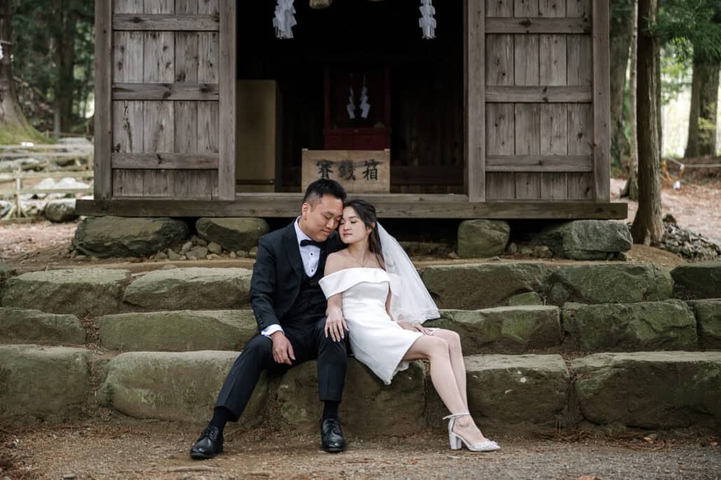 The couple sit close on the stone steps of a small wooden hall at Kawaguchi Asama Shrine, heads together and eyes closed, cedars on either side.