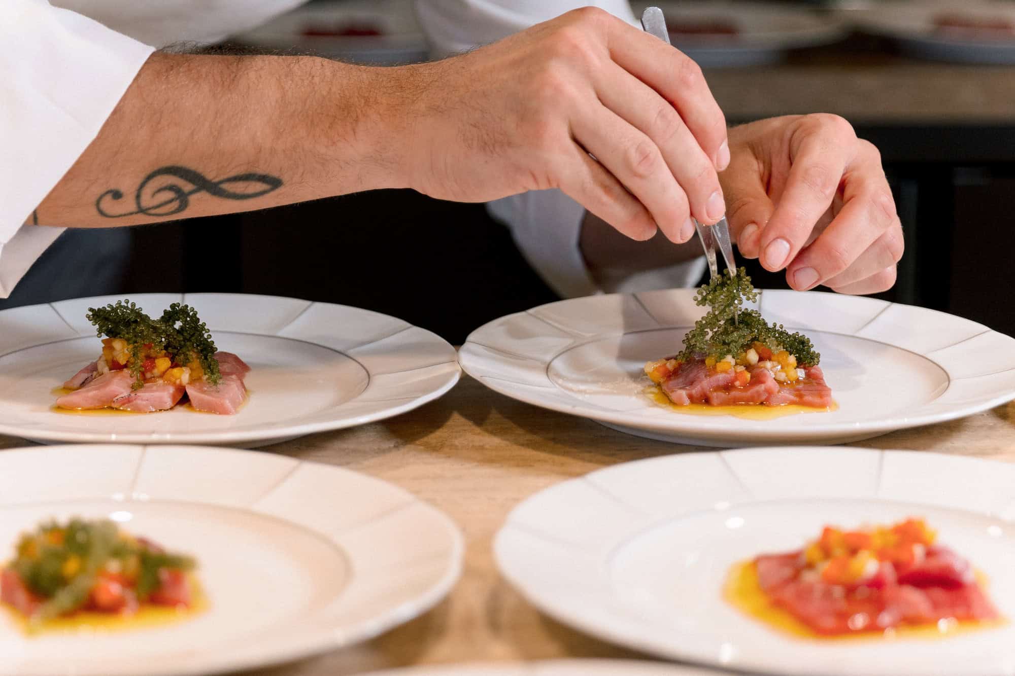 A chef uses tweezers to set umibudo, Okinawa's sea grapes, on tuna carpaccio across four white plates for a wedding lunch in Okinawa.
