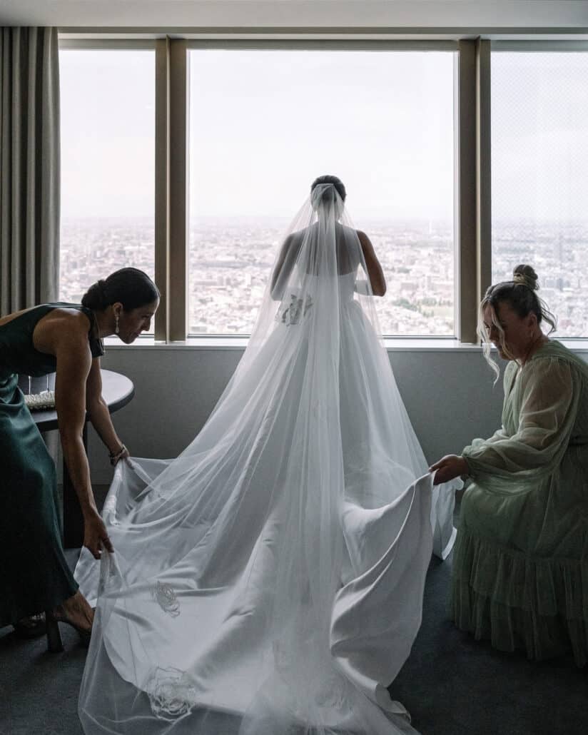 Two friends spread the bride's cathedral veil and train as she stands at the window over Tokyo, getting ready for her wedding.