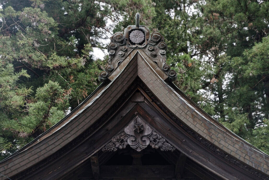 A cherry blossom crest sits at the peak of a roof gable at Kawaguchi Asama Shrine, ringed by curling cloud scrolls, with tall evergreens behind it.