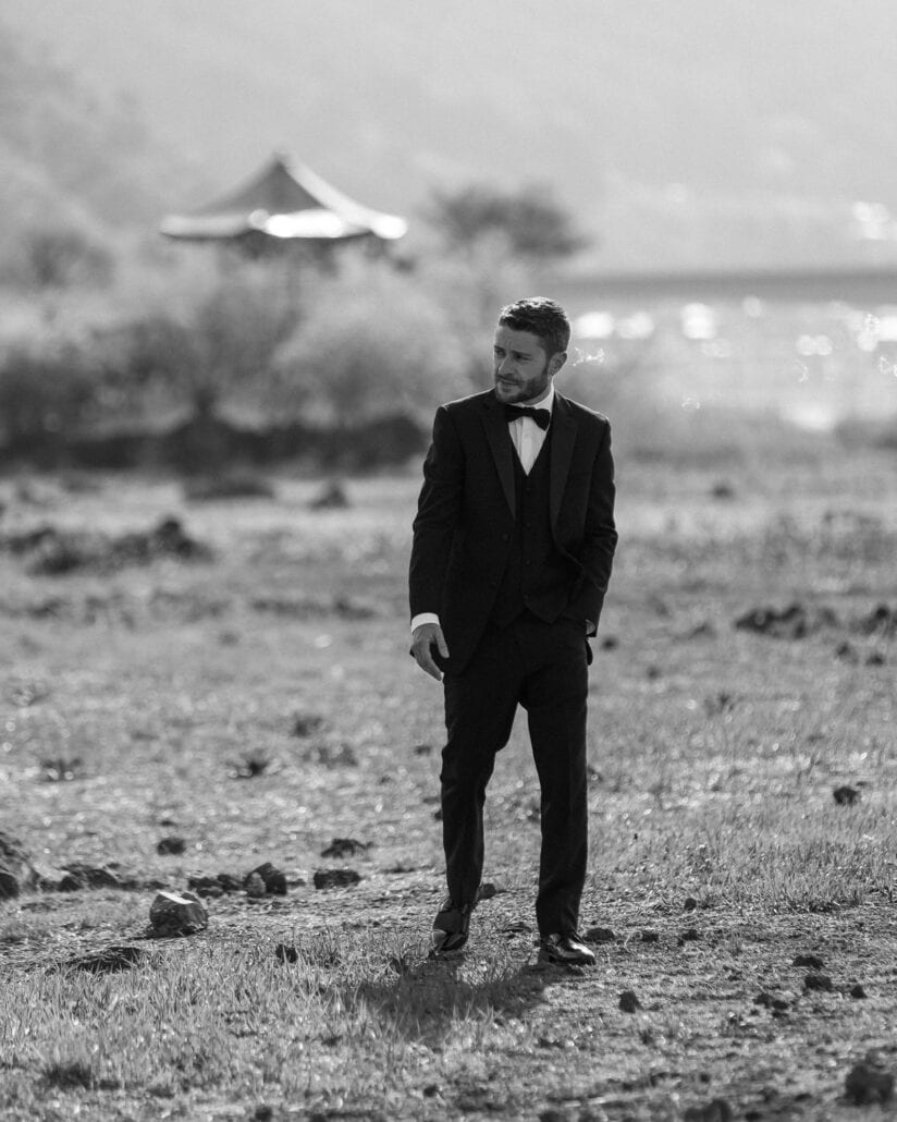 The groom in black tie stands on the lava-rock shore of Lake Kawaguchi, one hand in his pocket, a lakeside pavilion and bright water behind him.