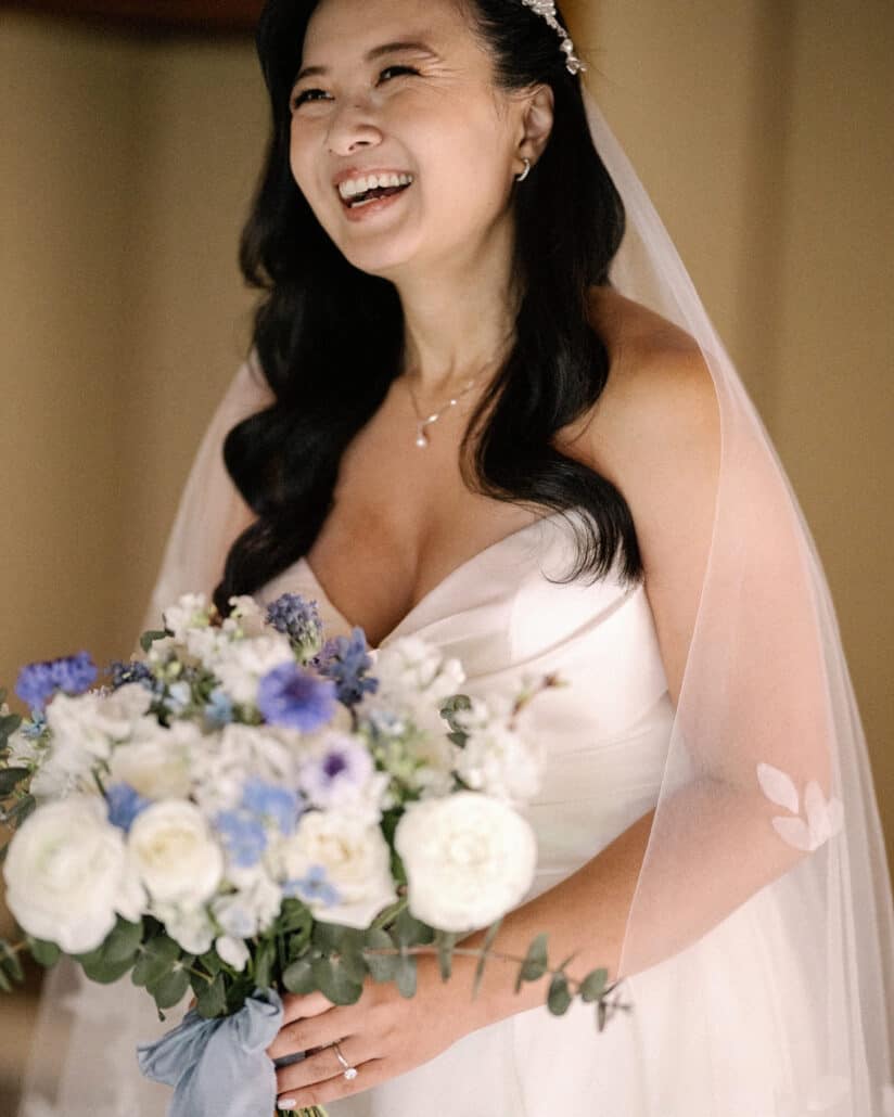 The bride laughs, holding her white roses and blue flowers, in her veil and a jewelled hairpiece before her destination wedding in Japan.