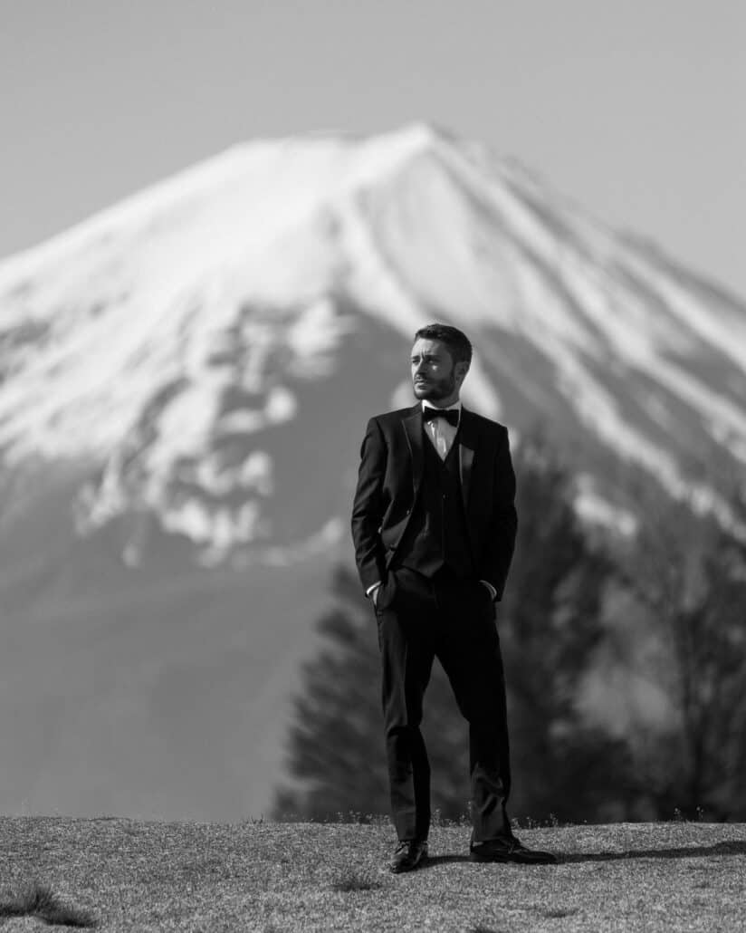 The groom in black tie stands with his hands in his pockets on the lawn at Lake Kawaguchi, Mt Fuji filling the sky behind him, in black and white.