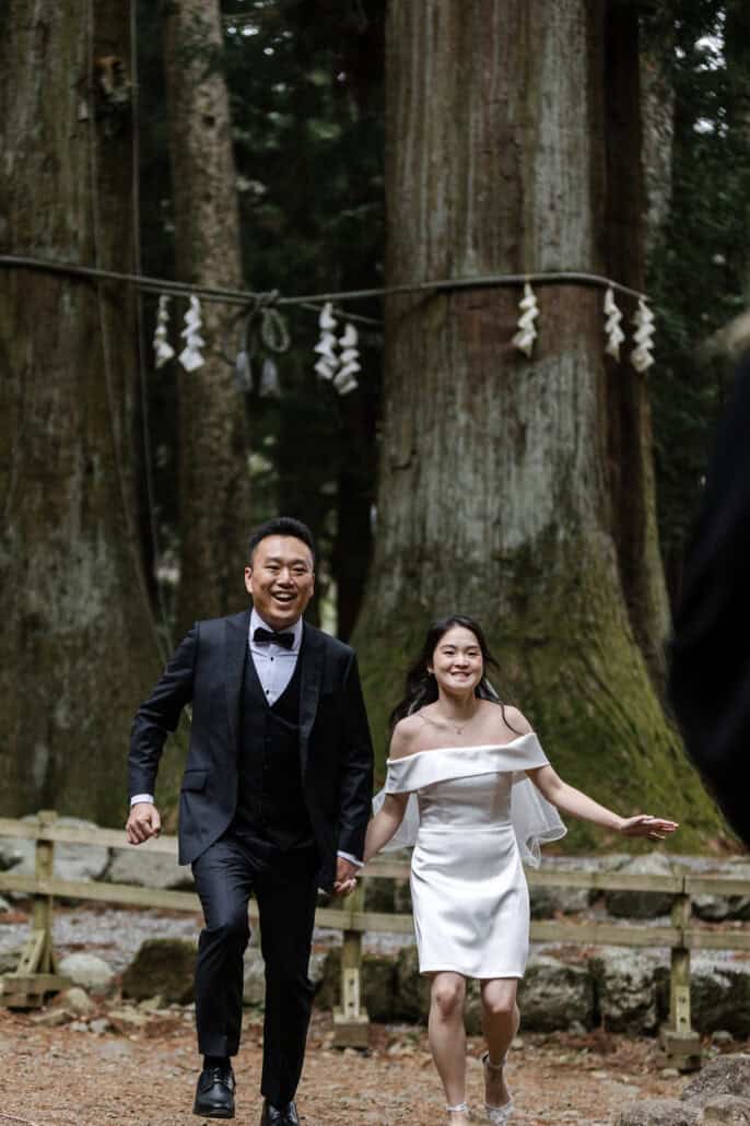 The bride and groom run toward the camera hand in hand and laughing, the roped matchmaking cedars of Kawaguchi Asama Shrine right behind them.