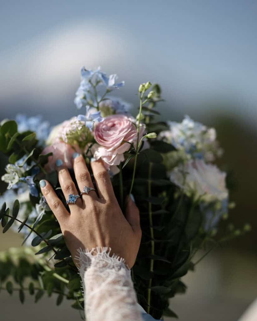 The bride's hand rests on her pink and blue bridal bouquet, showing her engagement ring with blue stones, a second ring and a lace cuff.