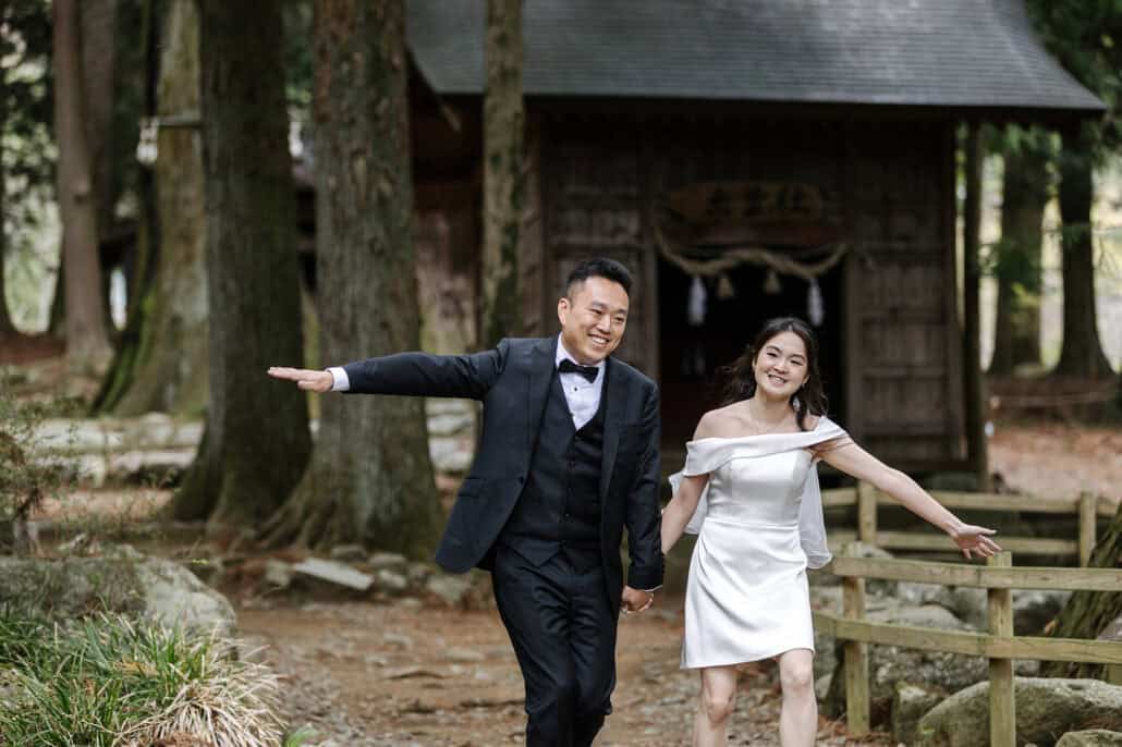 Smiling and hand in hand, the bride and groom stride out along a path from a small shrine hall, arms flung wide, the morning after their wedding.
