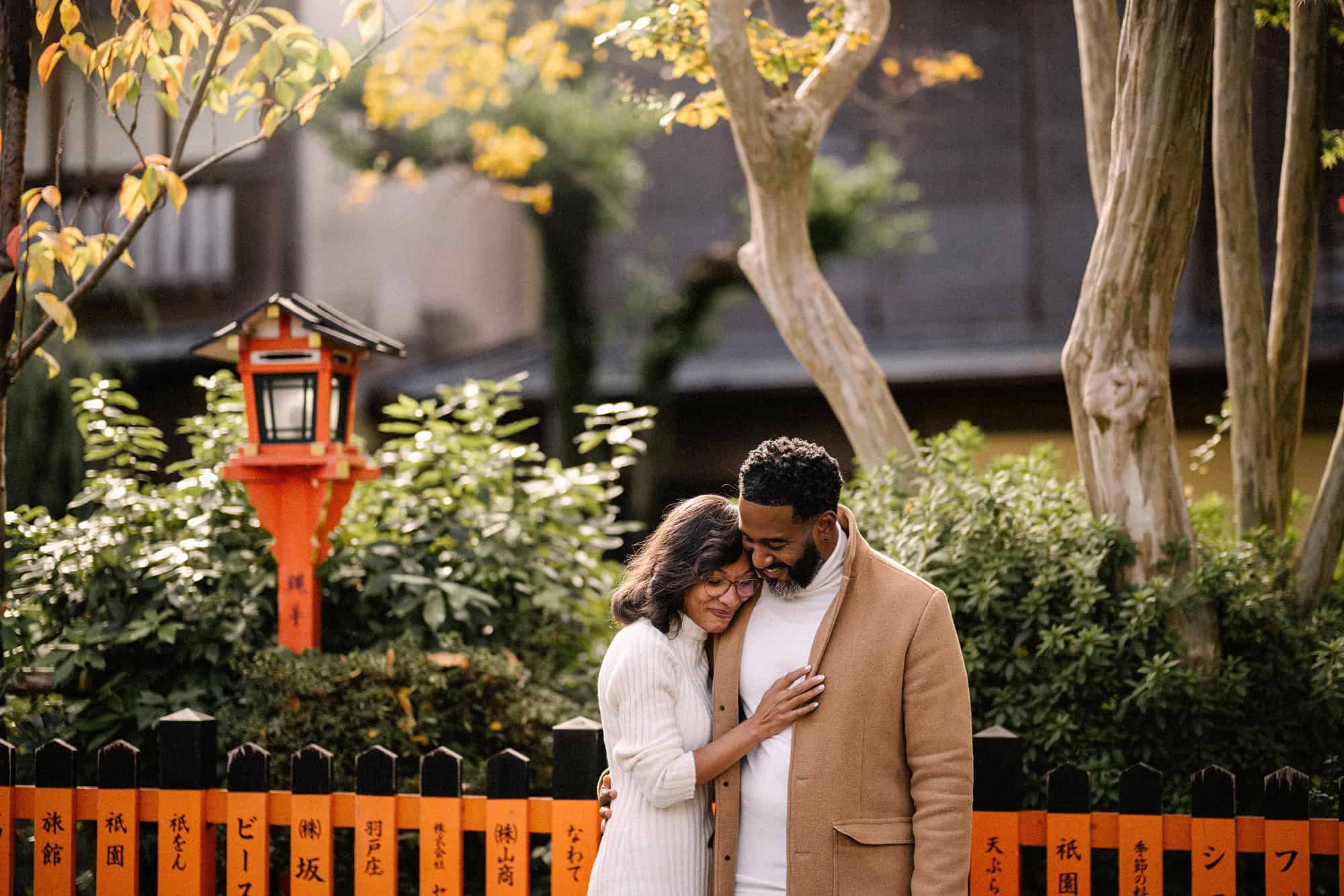 The couple hold each other by a vermilion shrine fence and lantern under yellow leaves in Gion Shirakawa, Kyoto, the day before their Japan elopement.