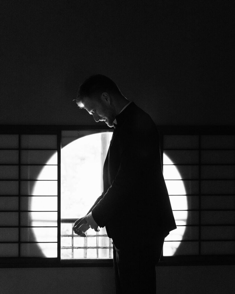 In black and white, the groom bows his head in silhouette against a round window and shoji screens before his ryokan wedding in Japan.