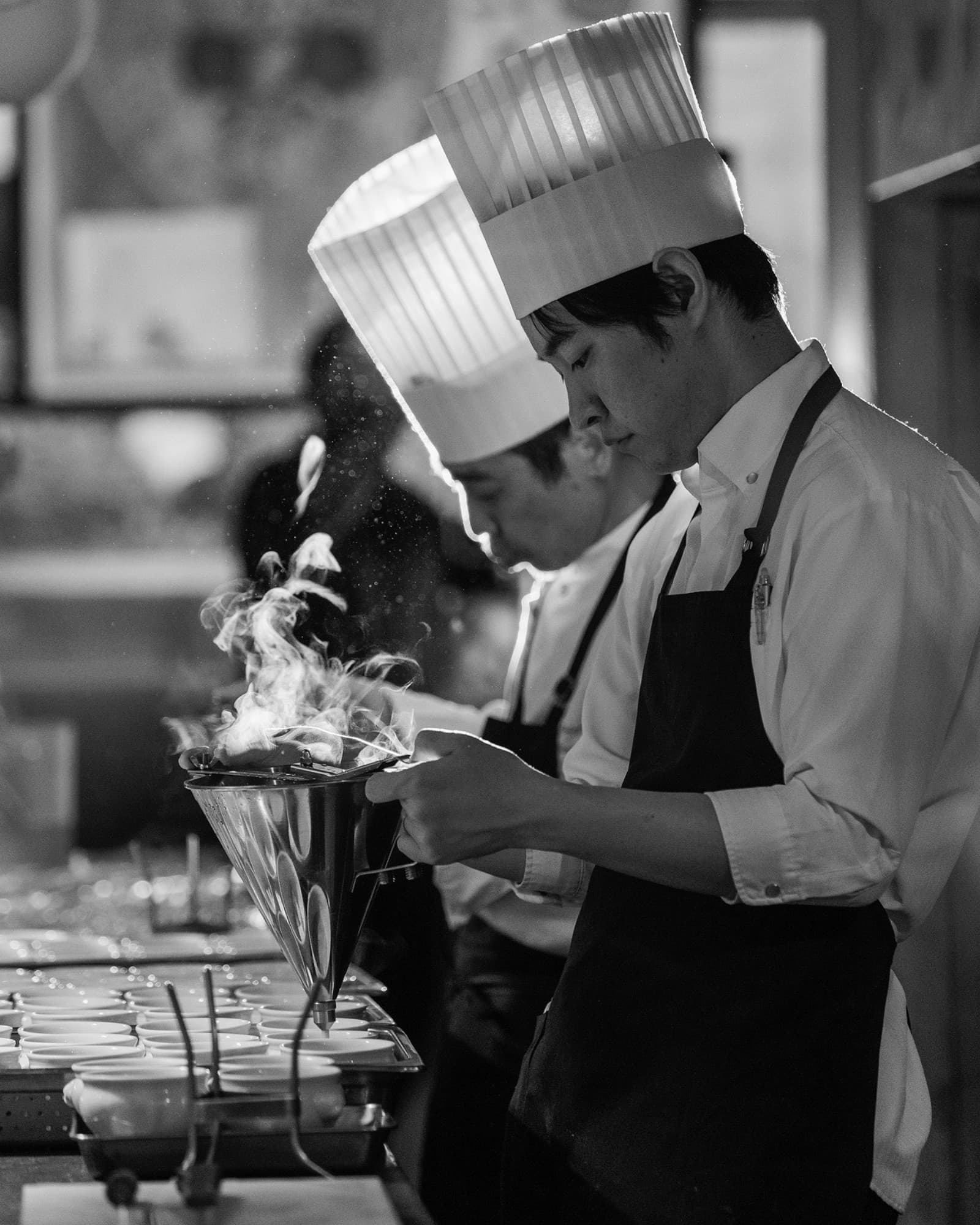 Two chefs in tall white hats fill rows of small bowls through a plume of smoke, a wedding dinner being plated in Hakone, in black and white.