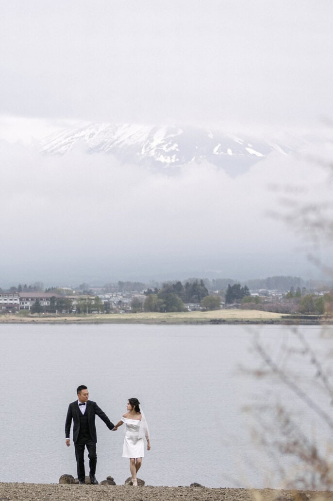 Hand in hand, the bride and groom walk the shore of Lake Kawaguchi toward the camera, Mt Fuji's snowcap showing through cloud across the water.