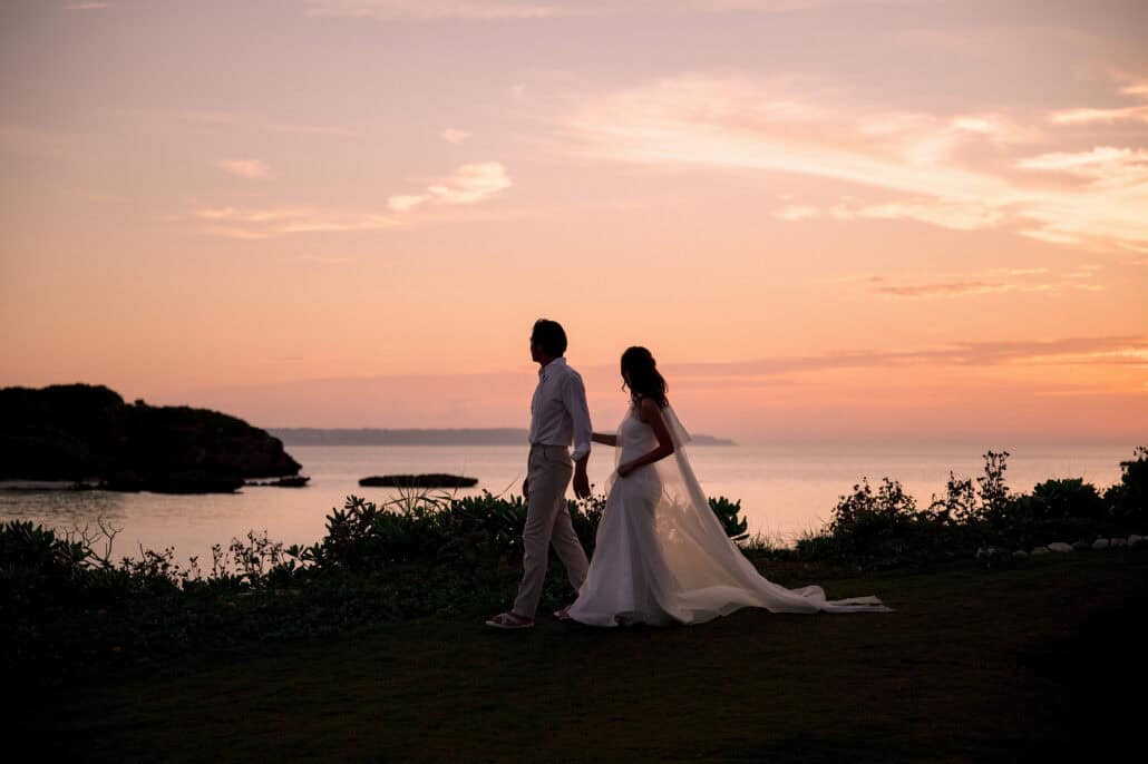 The bride and groom walk along a clifftop above the sea at sunset, her long veil trailing on the grass, on the evening of their wedding in Okinawa.