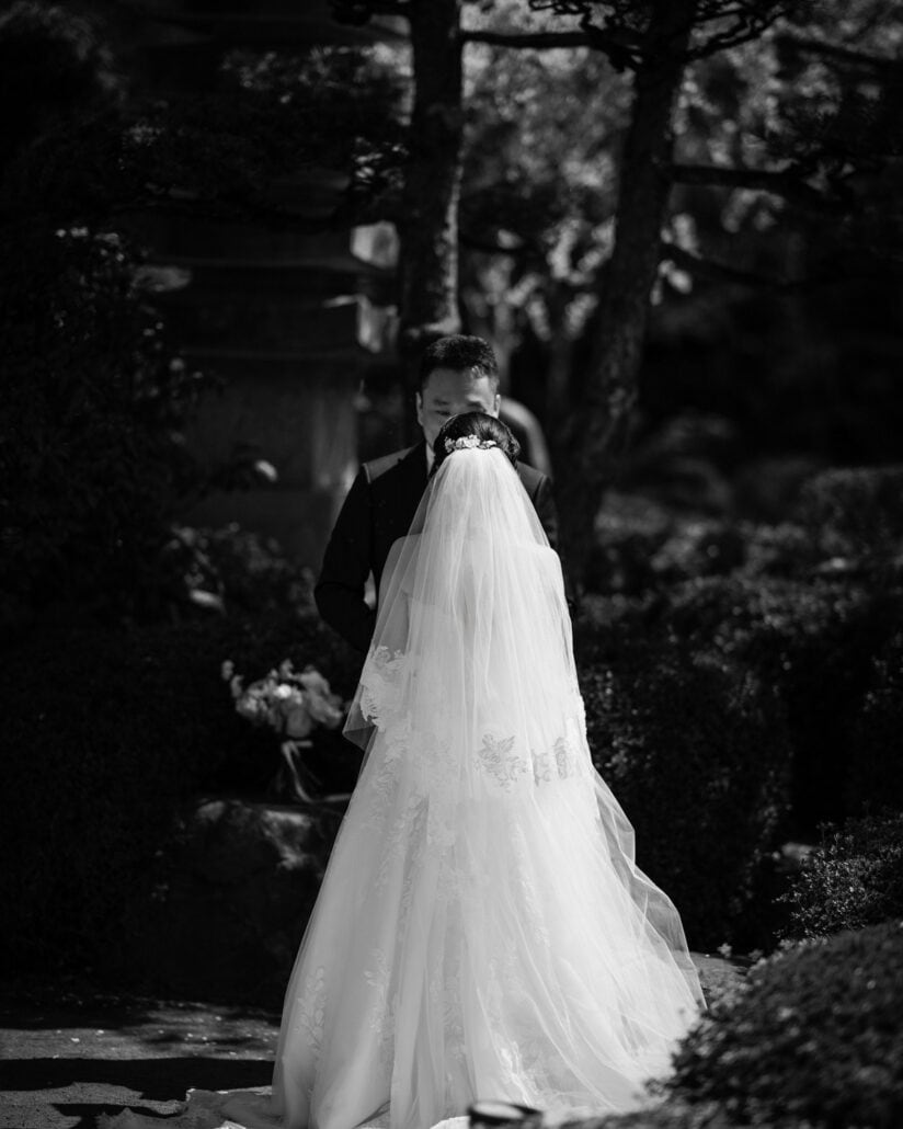 In black and white, the bride and groom meet at their first look in a shaded Japanese garden, her long veil falling to the ground.
