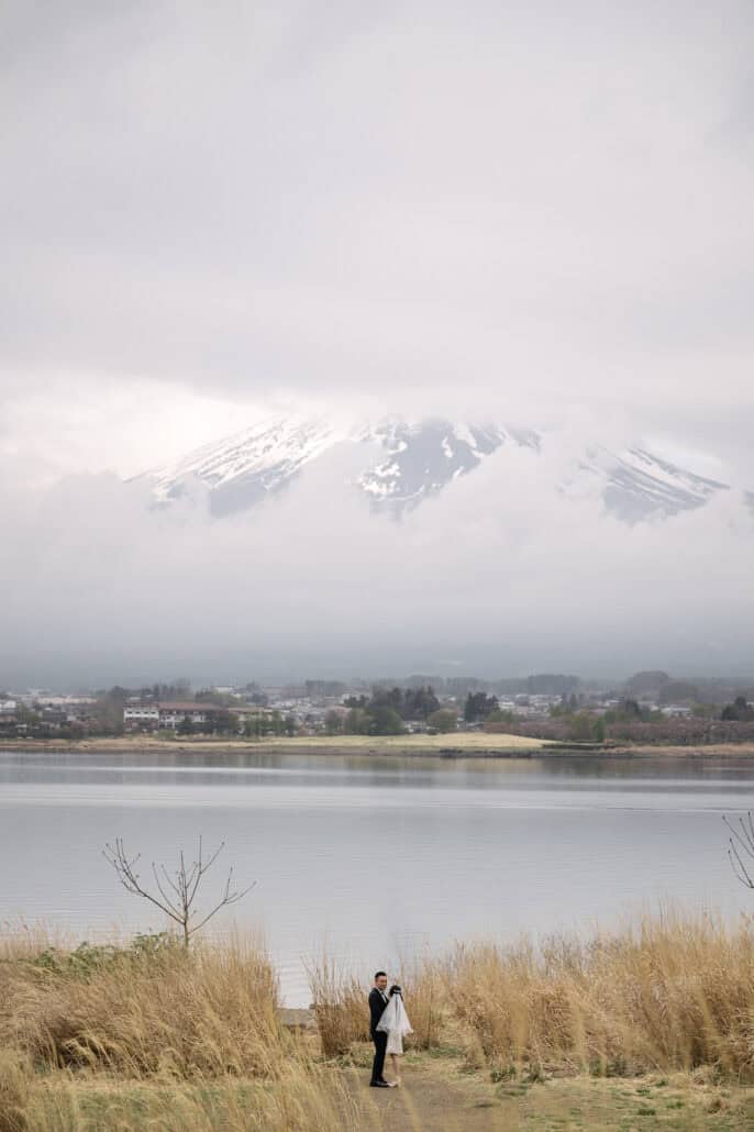 The couple stand tiny among tall golden grass at the edge of Lake Kawaguchi, the snowcap of Mt Fuji huge in the cloud above them.