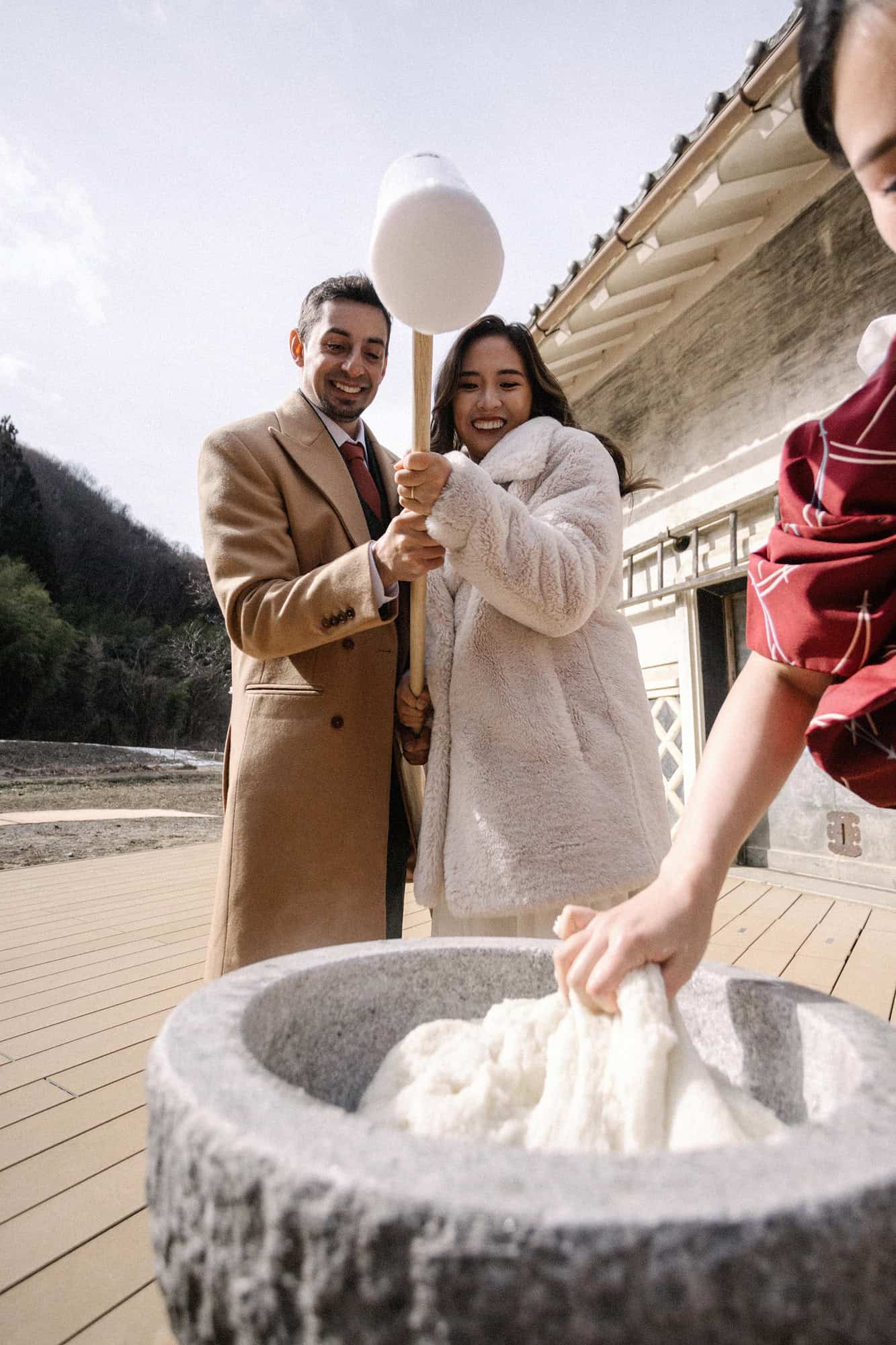 The bride and groom swing a wooden mallet together over a stone mortar for mochi pounding as a hand in a red kimono sleeve turns the rice.