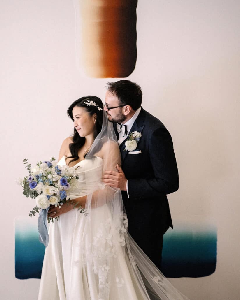 At Asaba ryokan, the groom kisses the bride's temple before a large painted artwork, her white and blue bouquet tied with a pale blue ribbon.